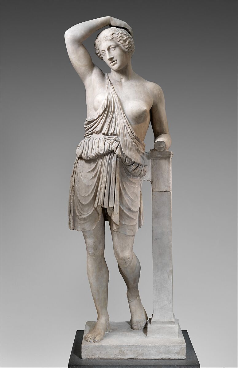 Statue de marbre : une femme debout, le bras droit relevé au-dessus de la tête, la tunique dégrafée à l'épaule, une blessure au sein droit.