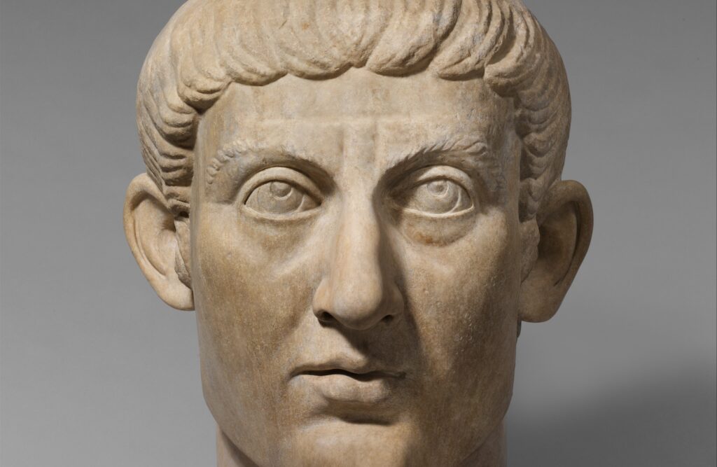 Tête colossale en marbre : un homme au visage allongé et glabre, les cheveux courts et soignés, le regard porté vers le haut.