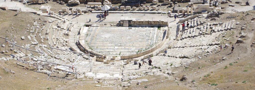 Vue plongeante sur les gradins de pierre d'un théâtre antique en hémicycle, avec l'orchestra circulaire au centre.