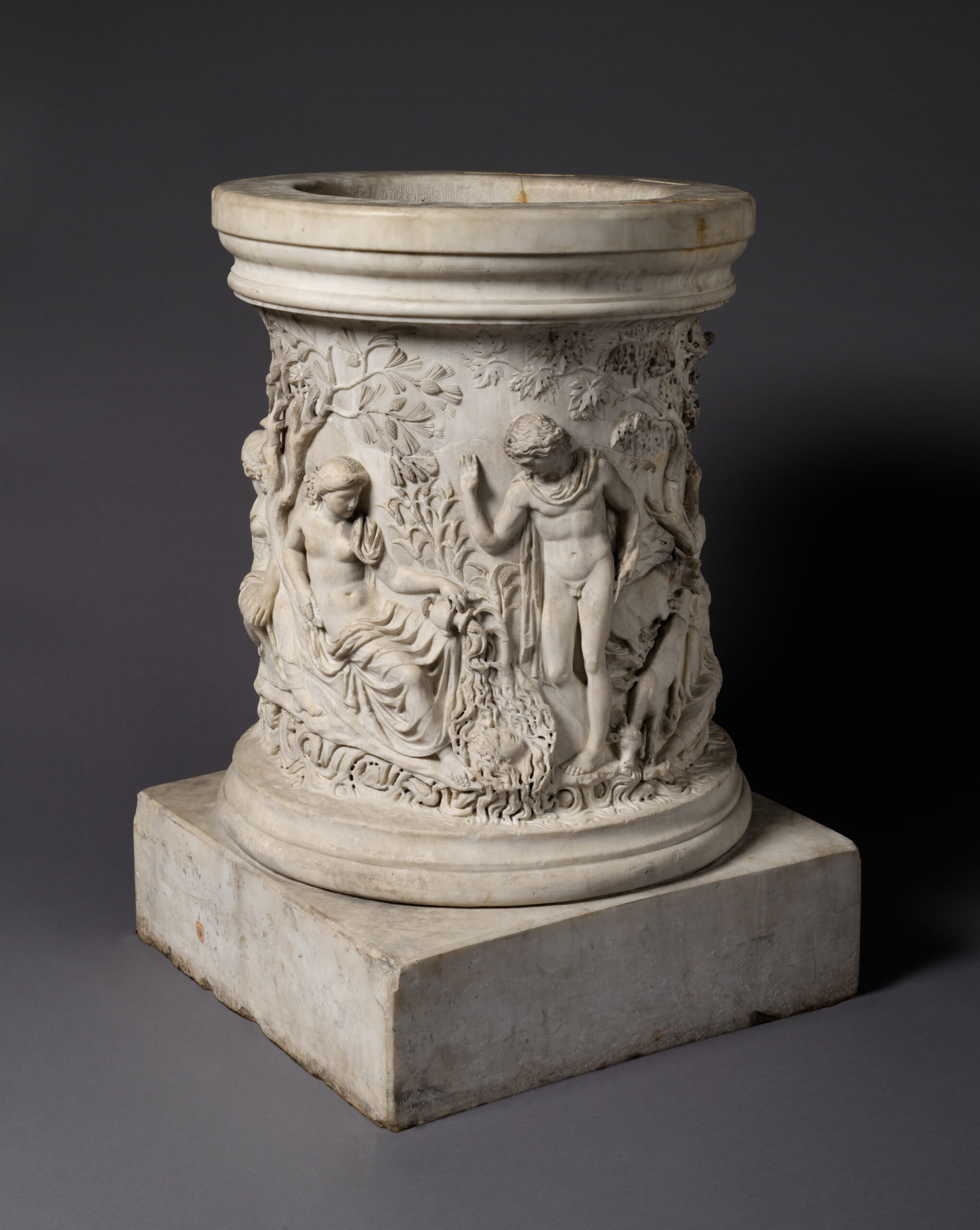Margelle de puits en marbre, sculptée en relief sur tout son pourtour : un jeune homme penché sur l'eau, une figure féminine, et un autre groupe où des nymphes entourent un jeune homme.