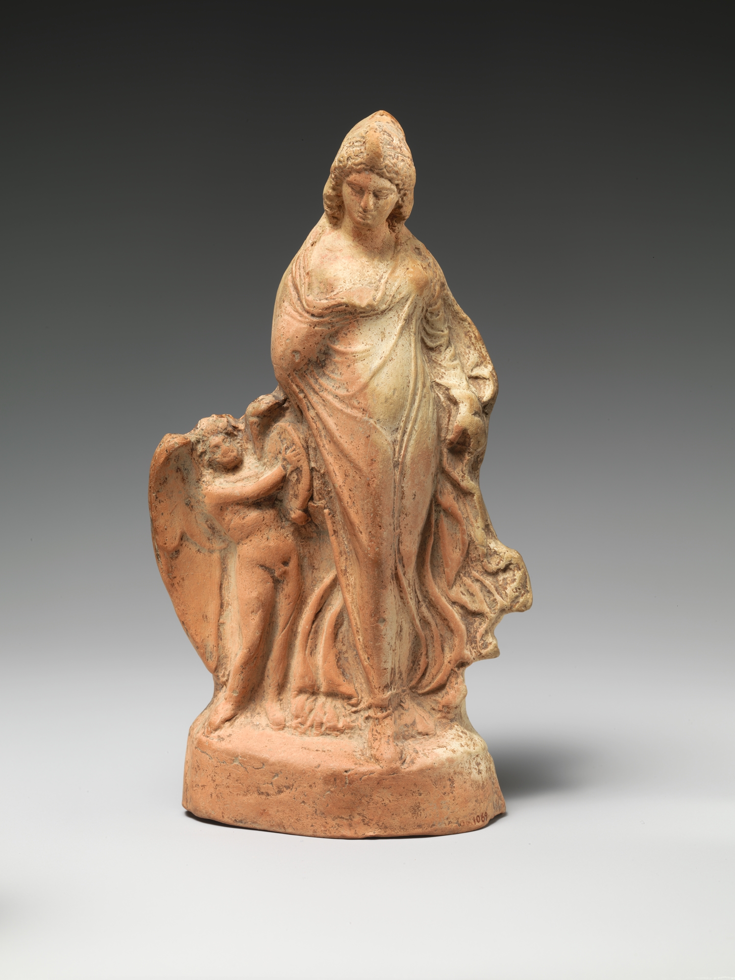 Statuette de terre cuite : une femme drapée en mouvement de danse, accompagnée d'une petite figure ailée.