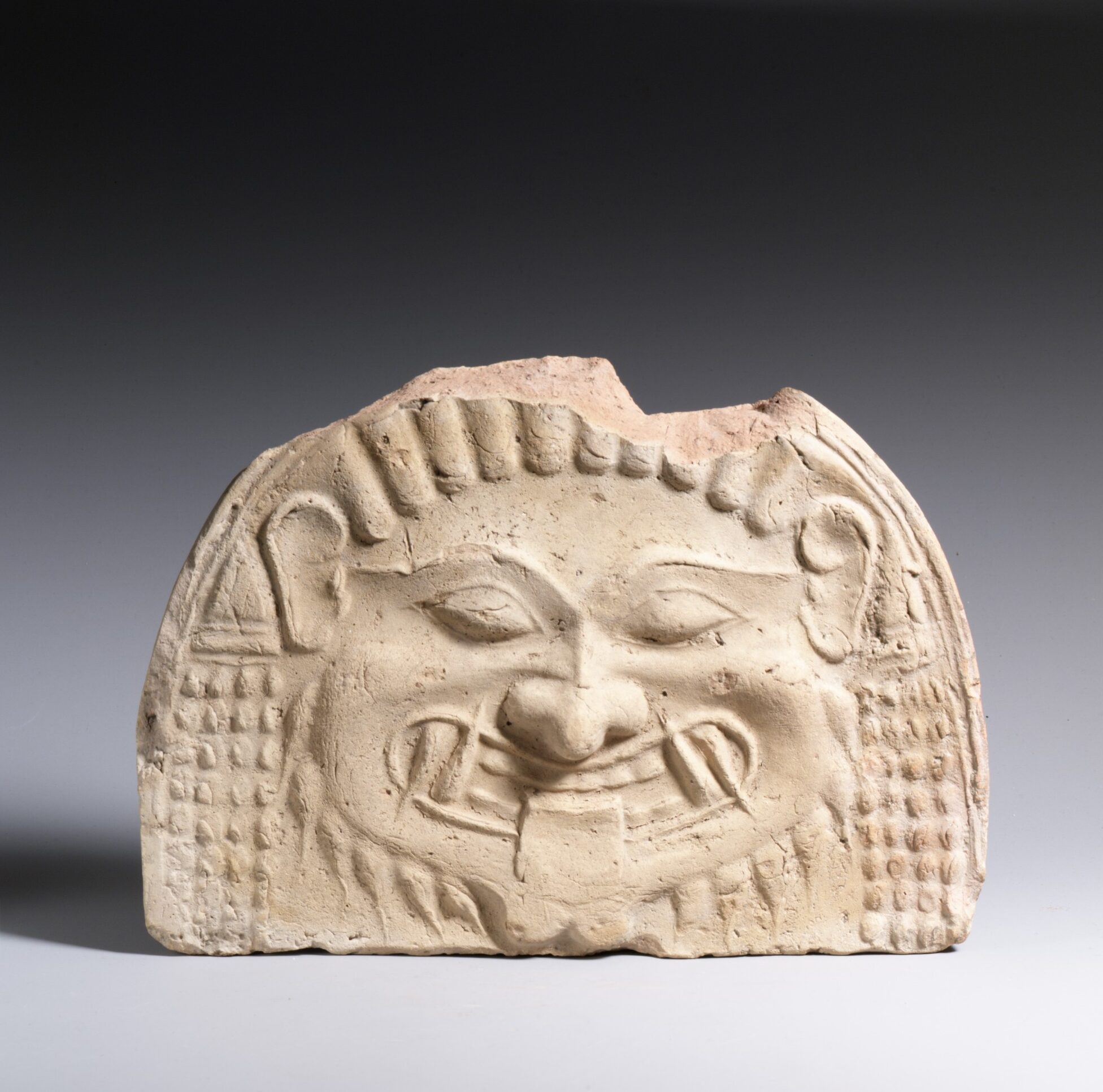 Tuile de terre cuite en forme de tête de Gorgone : yeux exorbités, bouche ouverte montrant des dents pointues, langue tirée.