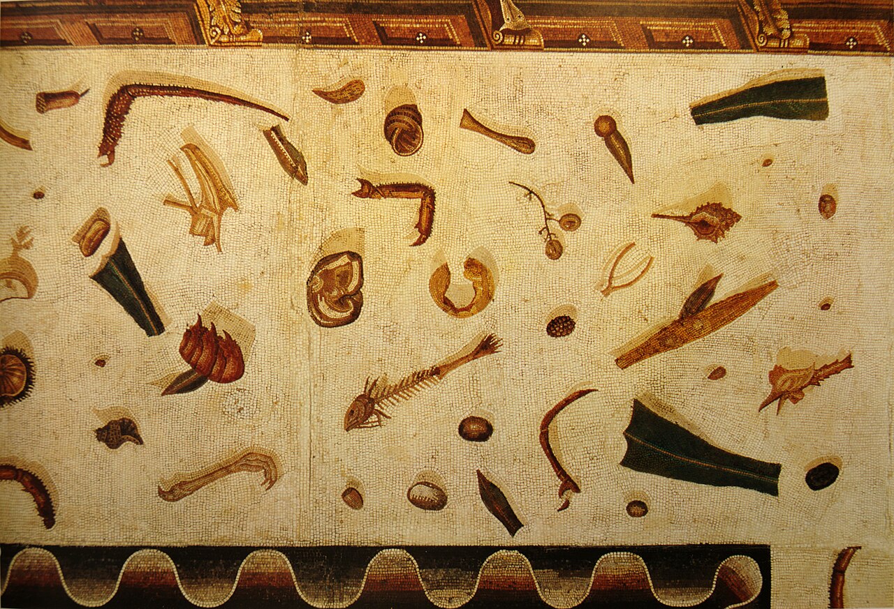 Sol de mosaïque à fond clair jonché de restes de repas représentés en trompe-l'œil, avec leurs ombres portées : arêtes de poisson, coquillages, pattes de volaille, coquilles de noix, une souris rongeant un fruit.