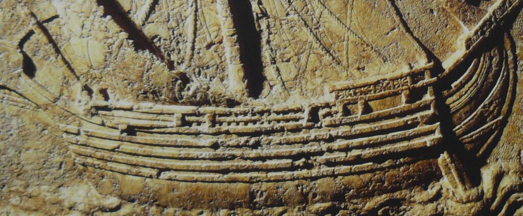 Bas-relief sculpté : un navire à voile aux extrémités relevées, avec son mât, ses vergues et ses cordages, voguant sur des vagues stylisées.