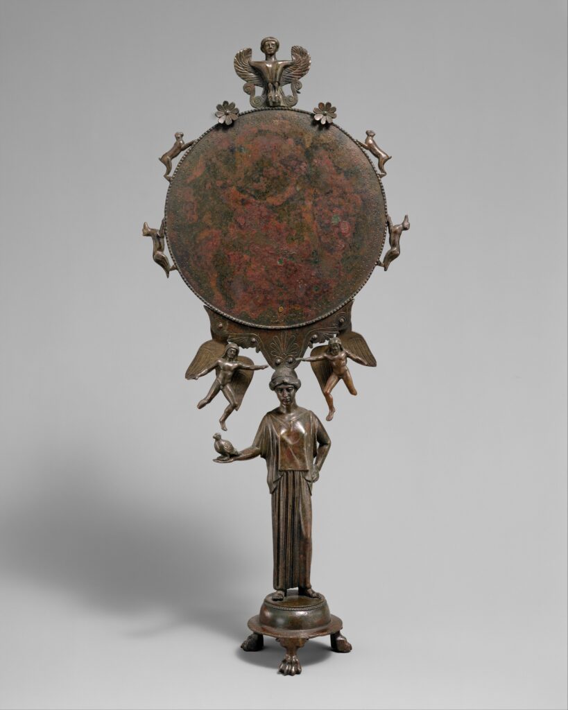 Miroir grec en bronze : le disque est porté par une statuette de femme drapée, surmontée d'une figure ailée et flanquée de petits personnages volants et d'animaux.