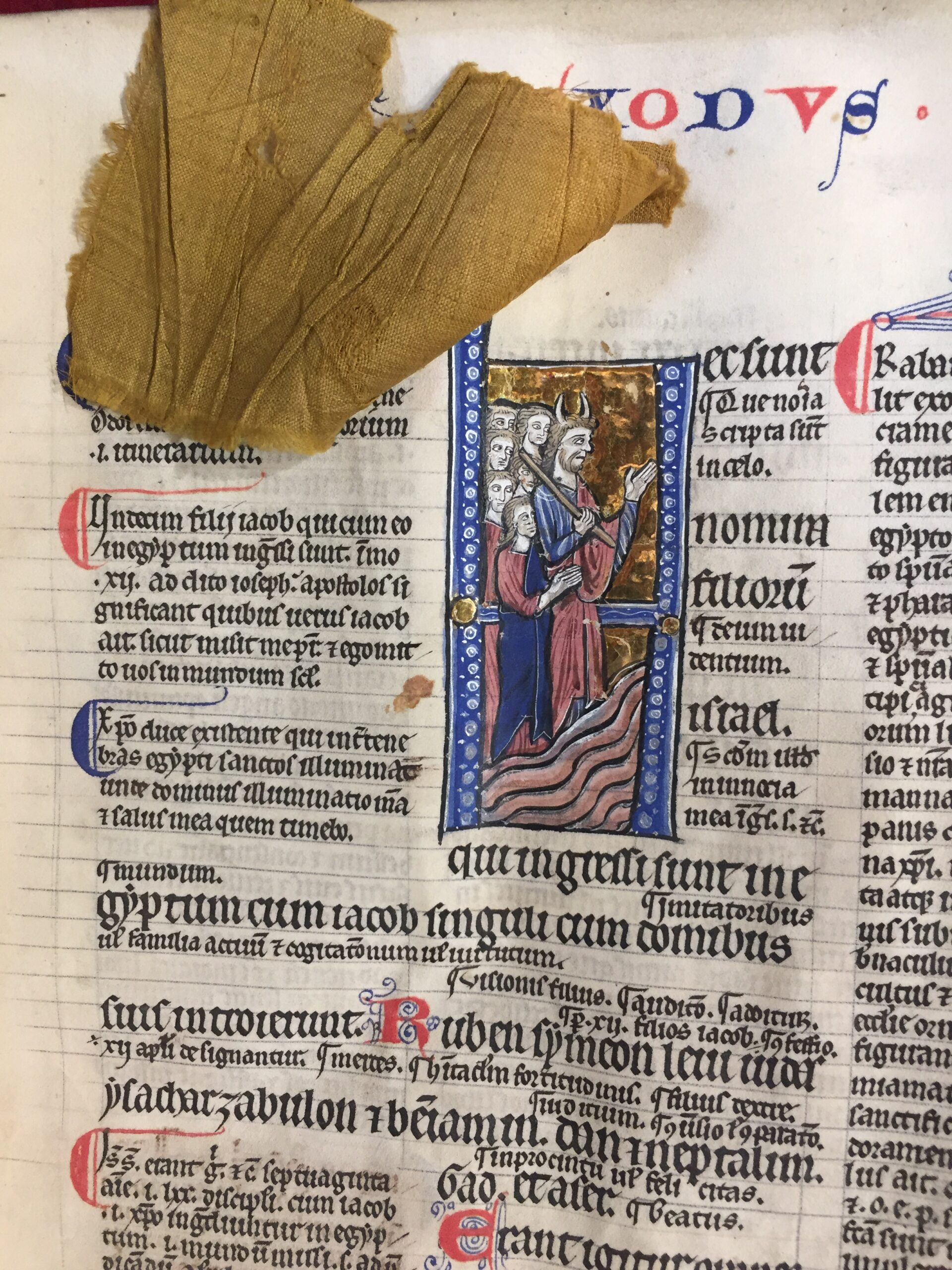 Main soulevant un fragment de tissu ocre cousu sur une page de manuscrit, découvrant une enluminure dorée.