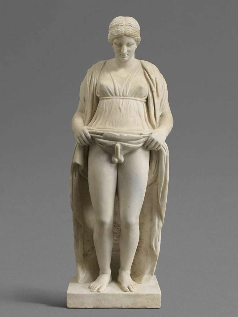 Statue en marbre d'une figure debout, vêtue d'une longue robe qu'elle soulève des deux mains, le visage baissé.