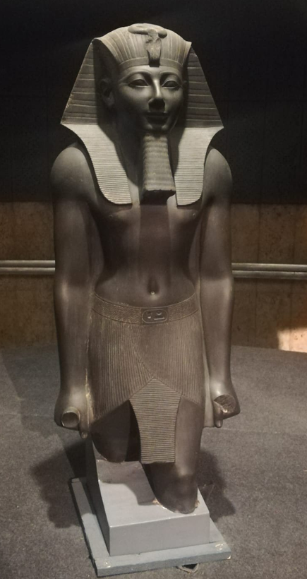 Statue en granite noir d'un roi debout, coiffé du némès, vêtu d'un pagne plissé, les bras le long du corps.